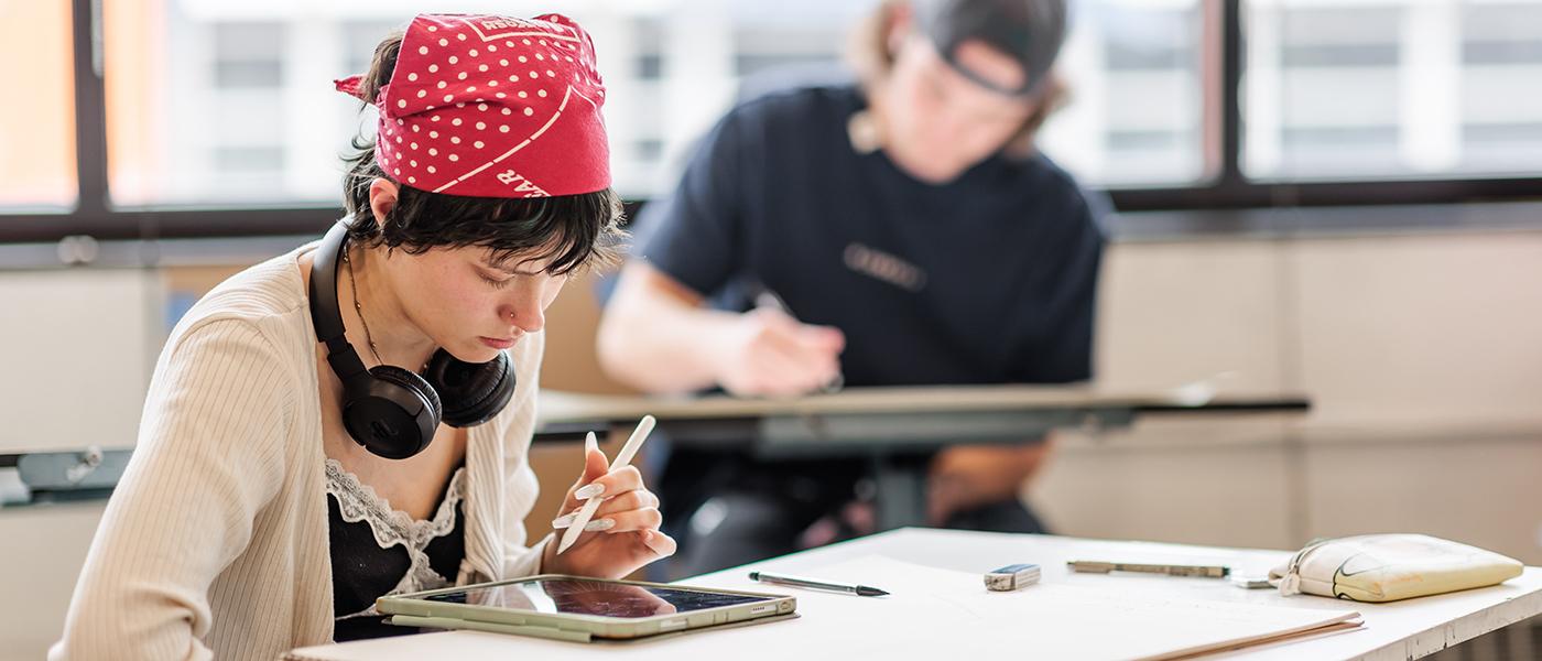 一名学生戴着红色圆点头巾，脖子上戴着耳机，用iPencil在iPad上画画.